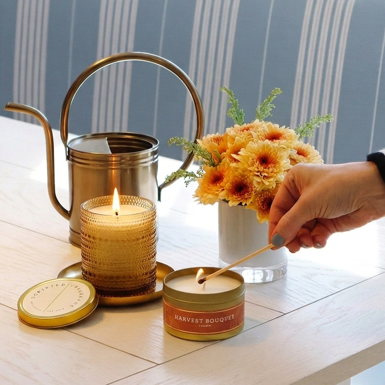 Lighting a Harvest Bouquet travel tin candle next to a lit Golden Grace soy candle on a dining table